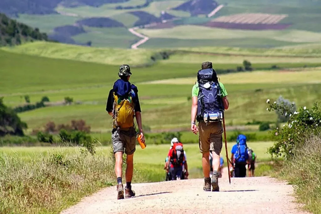Duas pessoas com mochilas caminhando em uma trilha de terra em meio a um cenário verde e montanhoso durante o Caminho de Santiago.