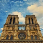 Vista de baixo para cima da fachada da Catedral de Notre-Dame em Paris, destacando as duas torres e a rosácea central contra um céu azul com nuvens.
