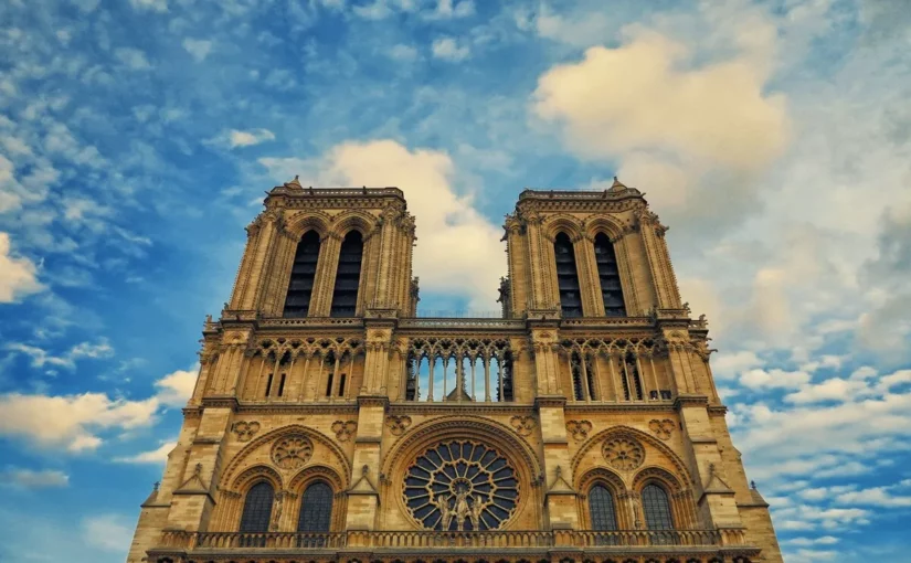 Vista de baixo para cima da fachada da Catedral de Notre-Dame em Paris, destacando as duas torres e a rosácea central contra um céu azul com nuvens.