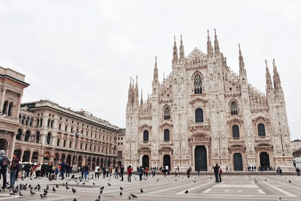 A Catedral de Milão (Duomo di Milano) na Europa, com sua fachada gótica em mármore branco e pináculos, em uma praça com pessoas e pombos.