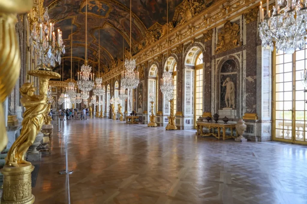 O interior ornamentado da Galeria dos Espelhos no Palácio de Versalhes, com seus lustres de cristal, estátuas douradas e teto pintado.