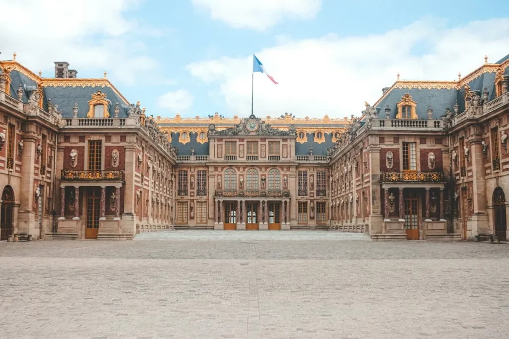 Fachada principal do Palácio de Versalhes, mostrando sua arquitetura clássica francesa, telhados dourados e um pátio de pedra.