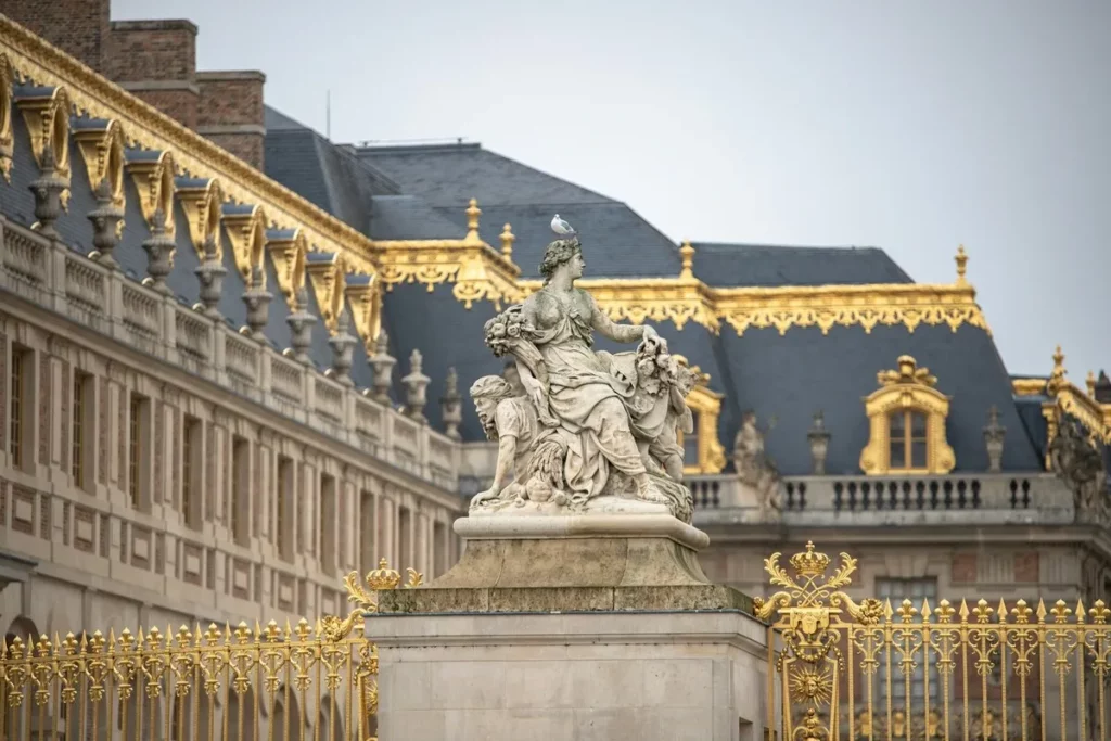 Estátua de mármore de uma figura mitológica em frente a um portão dourado e o telhado do Palácio de Versalhes, com detalhes em ouro.