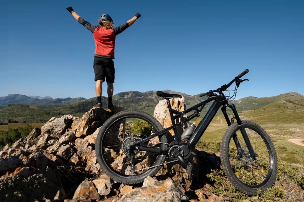 Ciclista com os braços erguidos em sinal de vitória no topo de uma montanha, ao lado de sua bicicleta de mountain bike.