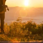 Uma pessoa com uma mochila de caminhada observando o nascer do sol em uma montanha, simbolizando uma aventura e exploração.