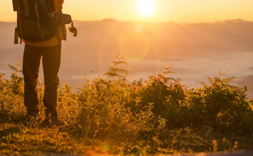 Uma pessoa com uma mochila de caminhada observando o nascer do sol em uma montanha, simbolizando uma aventura e exploração.