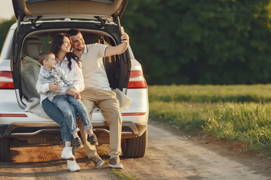 Família feliz sentada na mala de um carro, tirando uma selfie e curtindo um passeio de "bate-volta" no campo.