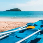 Caiaque azul com dois remos amarelos na areia da praia, com o mar azul e uma ilha ao fundo.