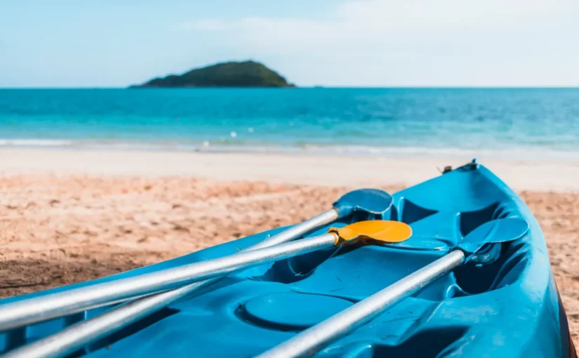 Caiaque azul com dois remos amarelos na areia da praia, com o mar azul e uma ilha ao fundo.