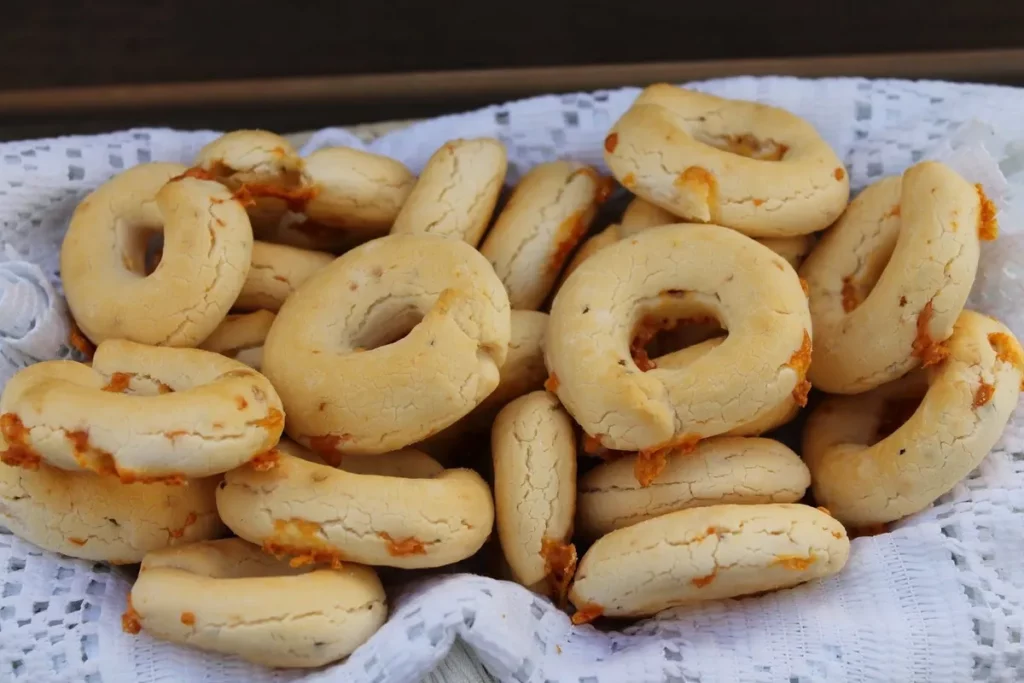 Uma bandeja com uma pilha de pães de queijo chipa, com a forma de anéis e ferraduras, em uma toalha de renda branca.