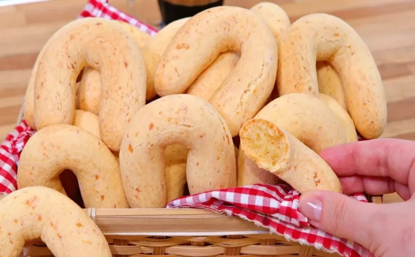 Uma cesta de vime forrada com uma toalha xadrez vermelha e branca, cheia de pães de queijo chipa em forma de ferradura, e uma mão pegando um deles.