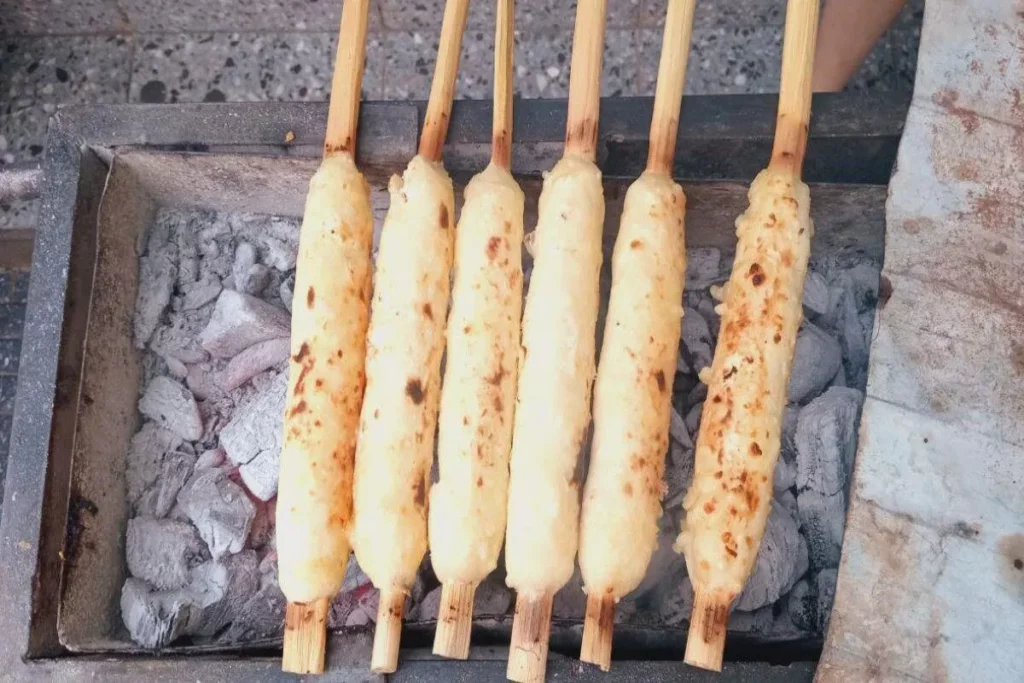 Seis espetos de comida típica da Bolívia sendo grelhados sobre carvão, em uma vista de cima.