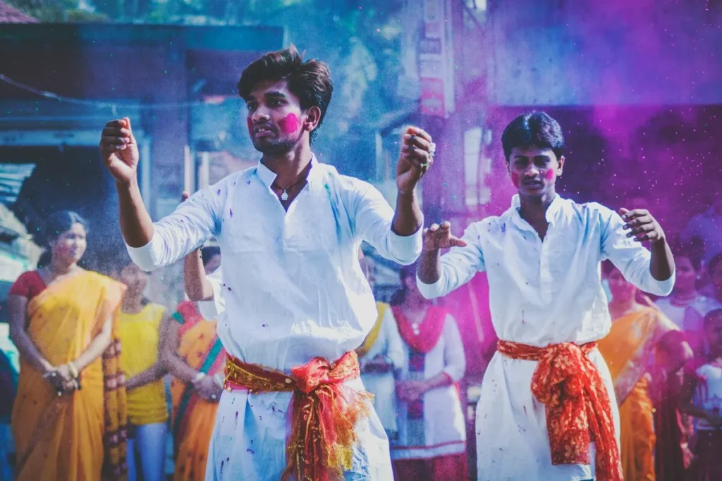 Homens dançando em um festival de cores, com vestimentas brancas e pó colorido no ar, representando a cultura e celebração.