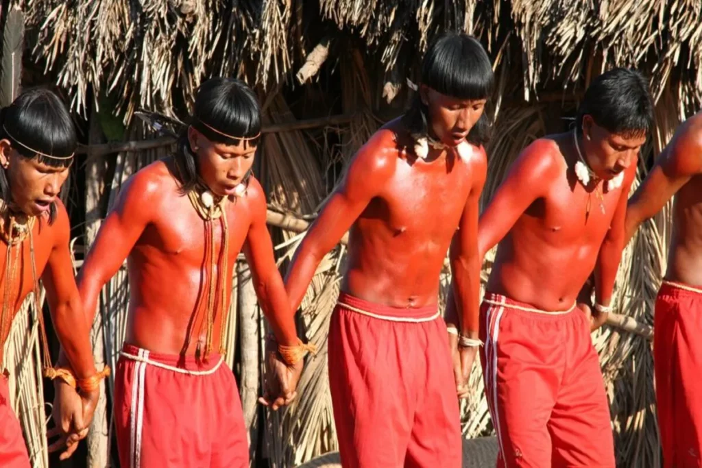 Grupo de pessoas com corpos pintados de vermelho, segurando as mãos em uma dança ou ritual, representando a cultura indígena.