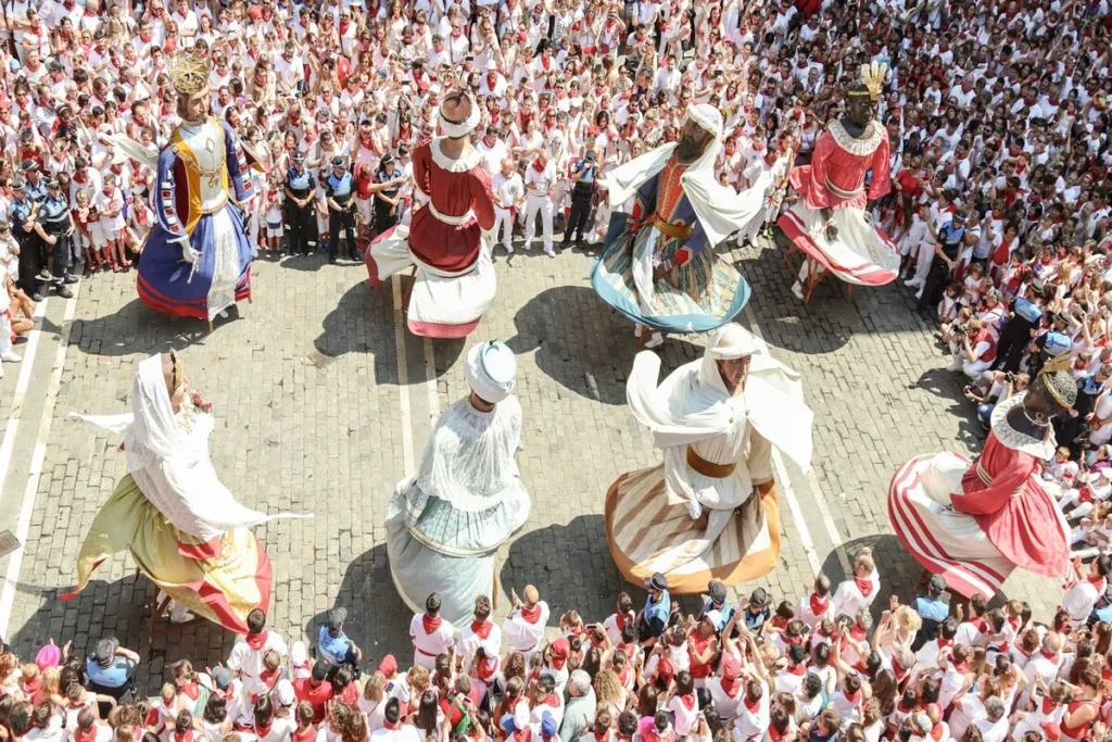 Visão aérea de um desfile cultural com figuras gigantes dançando em uma praça lotada de pessoas vestidas de branco e vermelho, representando a cultura espanhola.