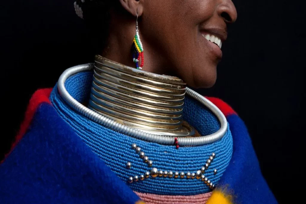 Mulher com colares tradicionais e intrincados, com contas coloridas e anéis de metal no pescoço, representando a cultura.