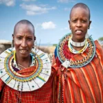 Retrato de duas mulheres Maasai usando cobertores vermelhos e colares de contas brancas e coloridas, representando a cultura e tradição.