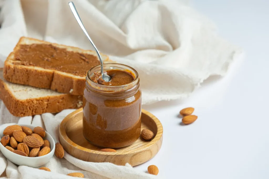 Um pote de vidro com doce de leite caseiro, com uma colher de metal dentro, ao lado de duas fatias de pão com doce de leite, amêndoas e um pano de linho branco.