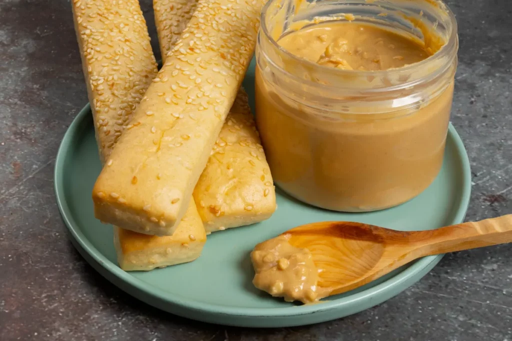 Um pote de vidro aberto com doce de leite cremoso ao lado de palitos de pão com gergelim em um prato verde, e uma colher de pau com um pouco de doce de leite na frente.