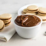 Um bowl branco cheio de doce de leite cremoso no centro, com dois alfajores à esquerda, um recheado e outro semi-recheado, e um prato com mais alfajores ao fundo, sobre uma superfície clara.