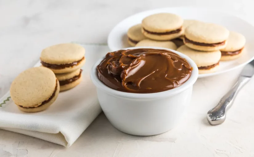 Um bowl branco cheio de doce de leite cremoso no centro, com dois alfajores à esquerda, um recheado e outro semi-recheado, e um prato com mais alfajores ao fundo, sobre uma superfície clara.