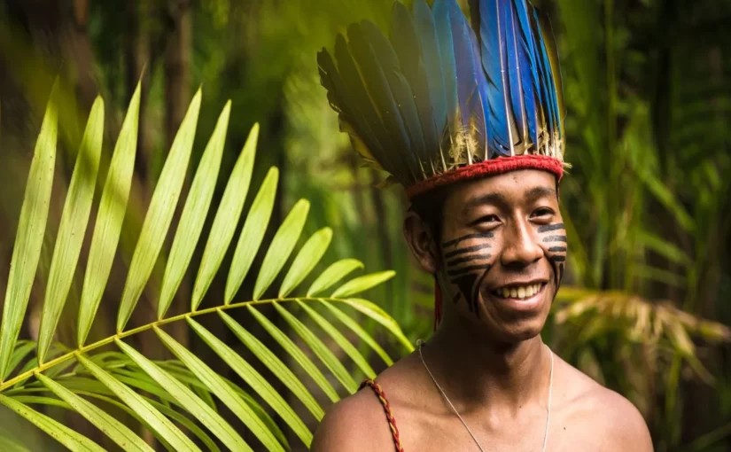 Retrato de um jovem indígena a sorrir, com um cocar de penas azuis e pinturas faciais tradicionais, num ambiente de floresta. A imagem representa a cultura e a história por trás das lendas indígenas.