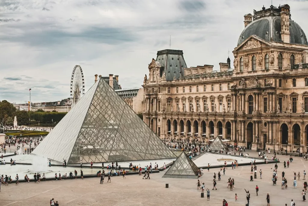 Vista exterior do Museu do Louvre, em Paris, com a icónica pirâmide de vidro no pátio central e a arquitetura clássica do palácio ao fundo. A imagem mostra a popularidade de um dos mais importantes museus de Paris.