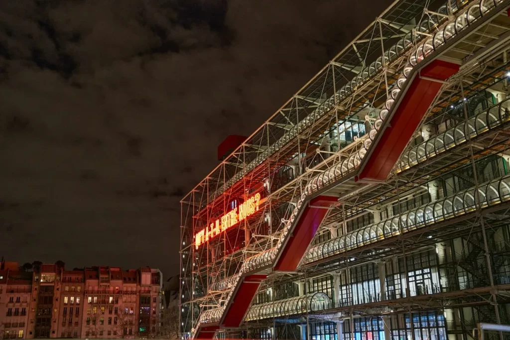 A fachada noturna do Centro Pompidou em Paris, com os seus tubos coloridos e escadas rolantes externas. A imagem destaca o design único de um dos mais famosos museus de Paris, com um letreiro de néon em vermelho a brilhar.