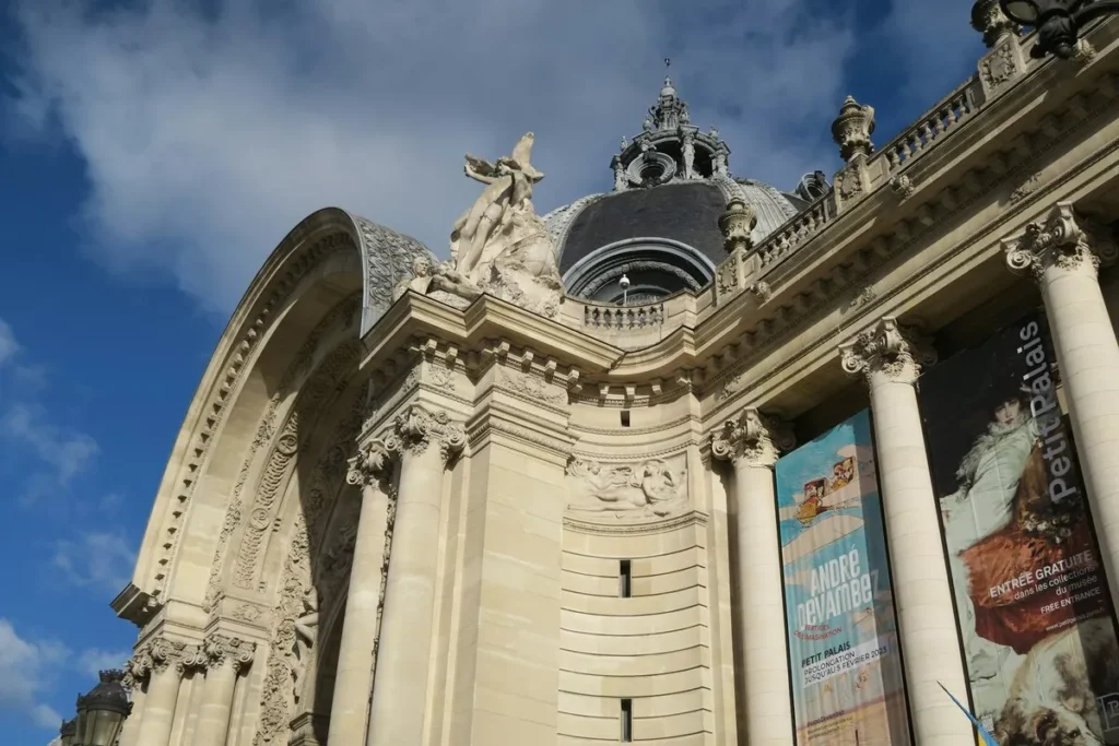 A fachada monumental do Grand Palais, em Paris, com a sua arquitetura clássica e esculturas, sob um céu azul. Um cartaz de exposição destaca a sua função como um dos importantes museus de Paris.