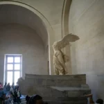 A icónica escultura "Vitória de Samotrácia", uma estátua de mármore de uma deusa alada sem cabeça, exposta no Museu do Louvre. A imagem mostra a sua grandiosidade, captando a admiração dos visitantes.