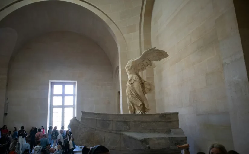 A icónica escultura "Vitória de Samotrácia", uma estátua de mármore de uma deusa alada sem cabeça, exposta no Museu do Louvre. A imagem mostra a sua grandiosidade, captando a admiração dos visitantes.