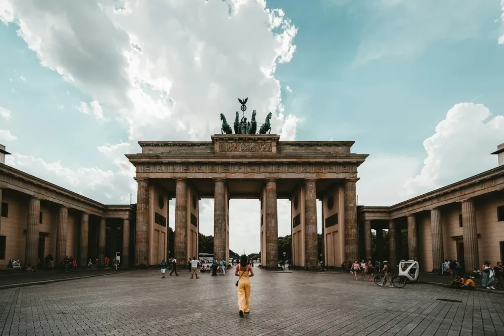 Vista da Porta de Brandemburgo, um dos principais monumentos de Berlim, com uma pessoa caminhando em primeiro plano e um céu azul com nuvens.