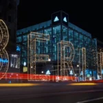 Vista noturna de um edifício de vidro da Adidas em Berlim, com uma iluminação de natal em formato da palavra "BERLIN" e rastros de luz de carros em movimento na rua.
