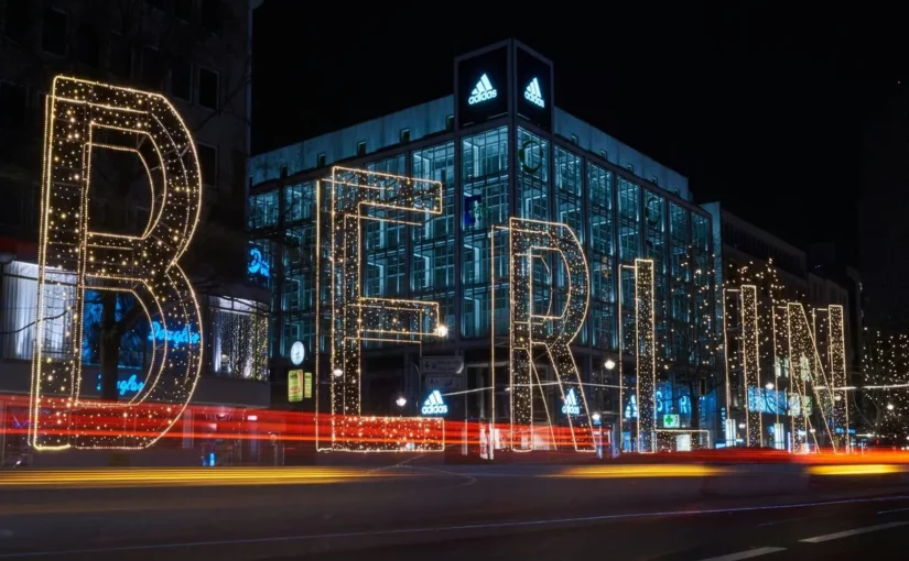 Vista noturna de um edifício de vidro da Adidas em Berlim, com uma iluminação de natal em formato da palavra "BERLIN" e rastros de luz de carros em movimento na rua.