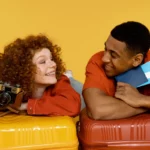Casal sorrindo e deitado sobre malas de viagem coloridas, segurando passagens e uma câmera, representando a alegria e a diversão de viajar juntos.