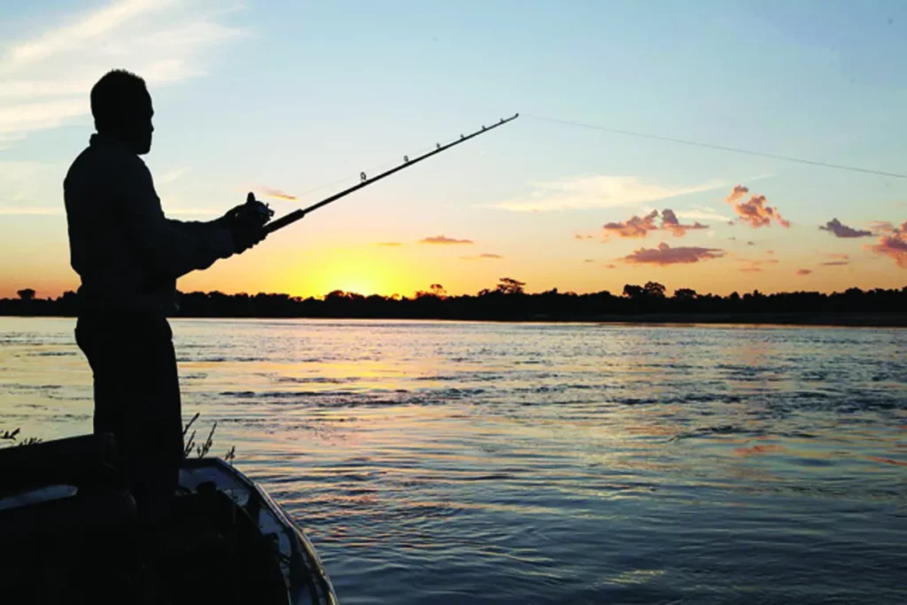 Silhueta de um pescador em um barco, pescando em um rio ao pôr do sol, praticando pesca esportiva.