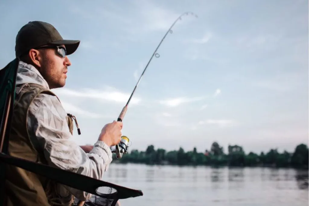 Homem com óculos de sol e boné sentado em uma cadeira de pesca, segurando a vara e olhando para o rio, praticando pesca esportiva.