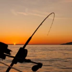 Vara de pesca com carretilha montada em um barco, em silhueta, com o sol se pondo no horizonte do oceano.