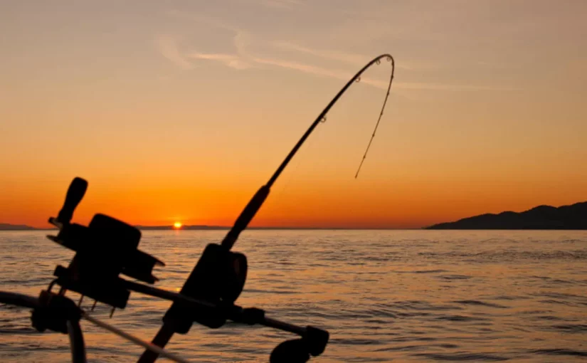 Vara de pesca com carretilha montada em um barco, em silhueta, com o sol se pondo no horizonte do oceano.