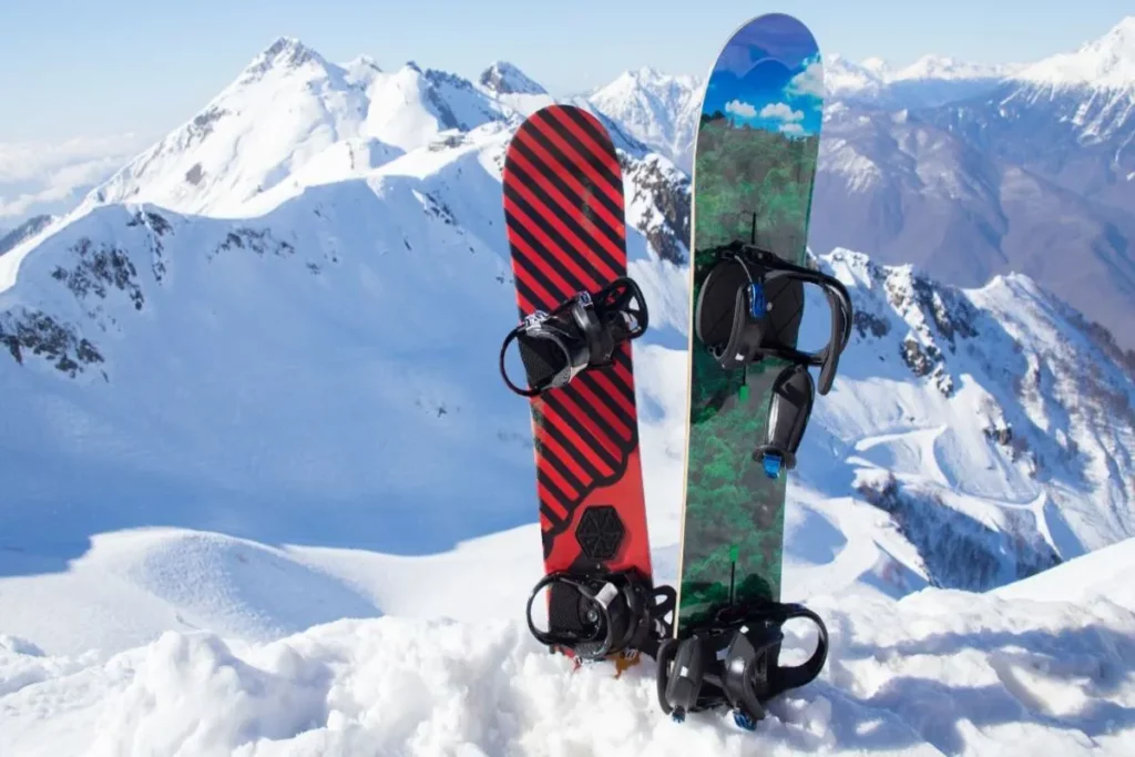 Duas pranchas de snowboard, uma vermelha e preta e outra verde e azul, em pé na neve, com uma paisagem de montanhas ao fundo.