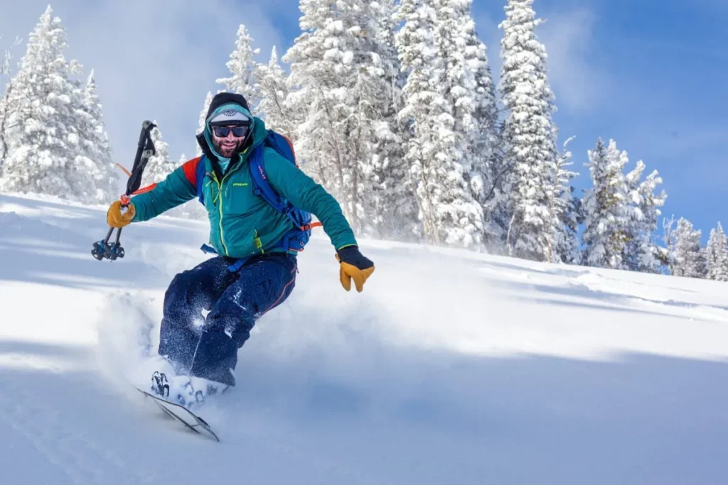 Homem sorridente em roupas de inverno praticando snowboard em uma montanha com neve, com árvores ao fundo.