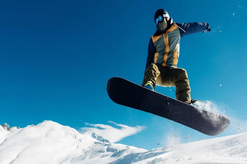 Snowboarder em manobra de salto no ar, com montanhas nevadas e céu azul ao fundo, praticando esporte radical.