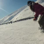 Mulher com gorro e jaqueta marrom e roxa praticando snowboard em uma montanha com neve, sob um céu ensolarado.
