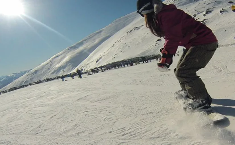 Mulher com gorro e jaqueta marrom e roxa praticando snowboard em uma montanha com neve, sob um céu ensolarado.