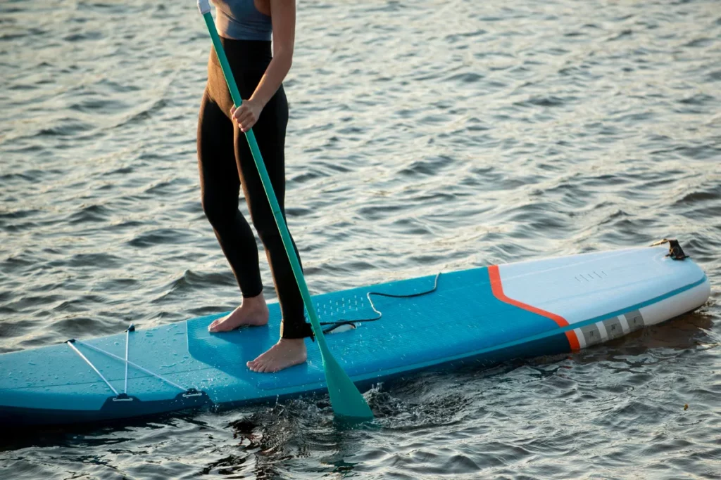 Uma pessoa em pé em uma prancha de stand up paddle, segurando um remo, praticando o esporte na água.