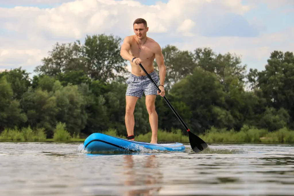 Um homem jovem e atlético praticando stand up paddle em um lago, com árvores ao fundo.