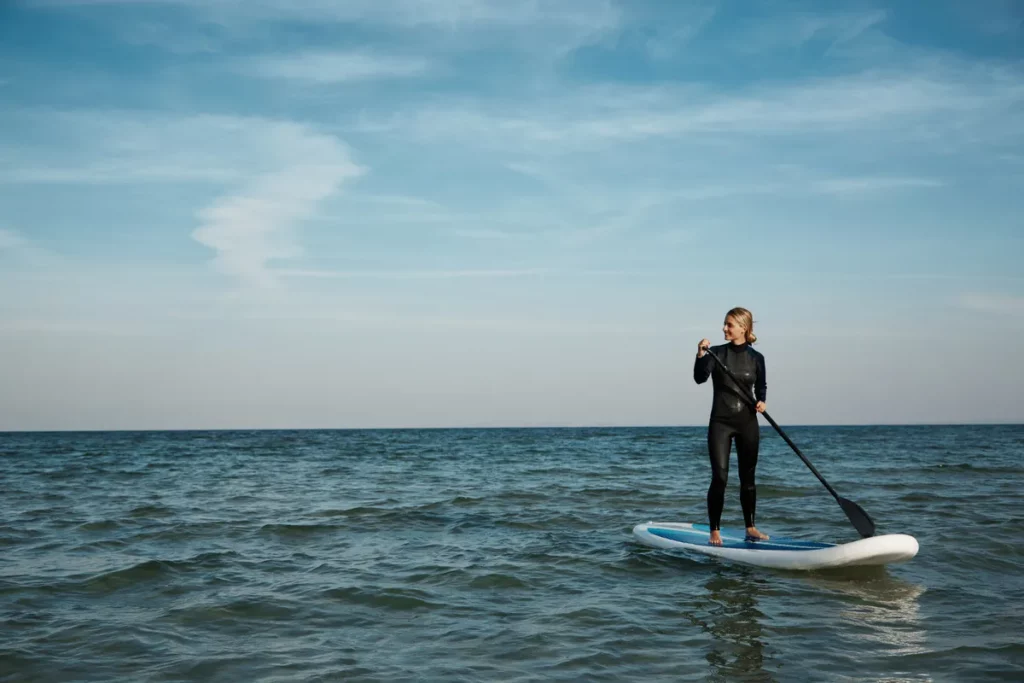 Mulher praticando stand up paddle em mar aberto, usando uma roupa de mergulho preta e segurando um remo.