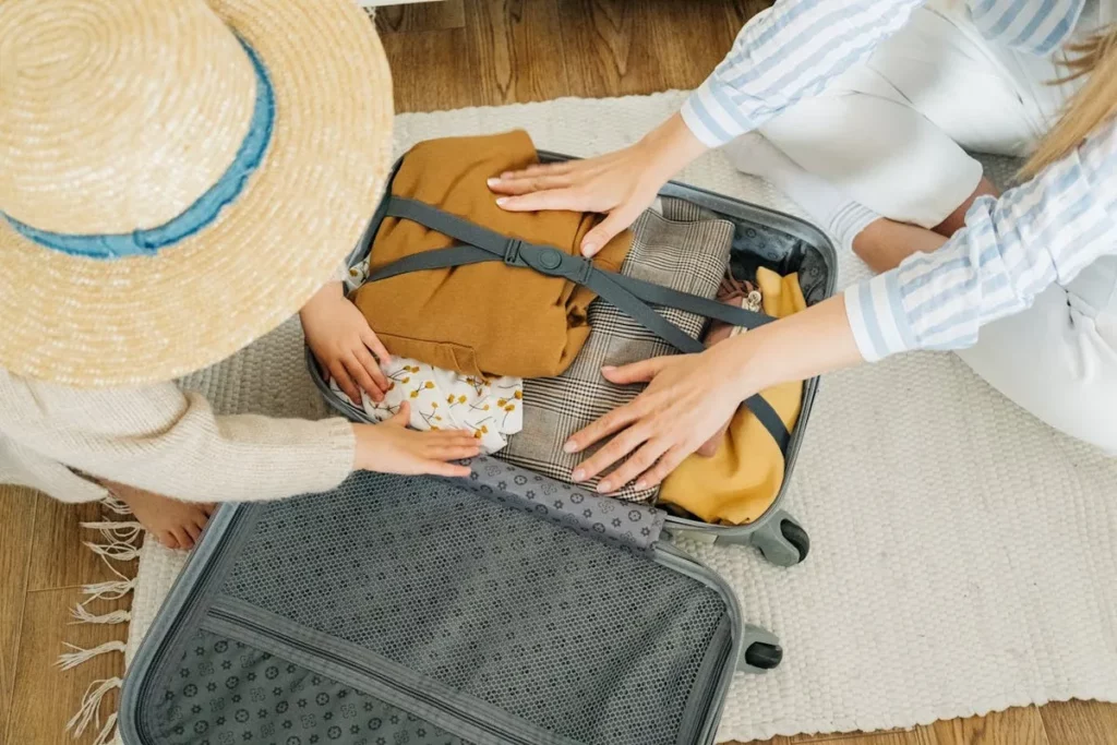 Uma mãe e uma criança pequenas estão arrumando a mala de viagem aberta no chão de madeira, em uma cena que mostra a preparação para uma viagem em família.