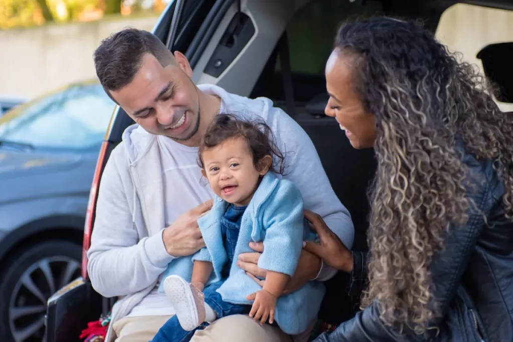 Um pai e uma mãe sorrindo, com o pai segurando um bebê que sorri para a câmera, enquanto estão sentados na parte de trás de um carro.