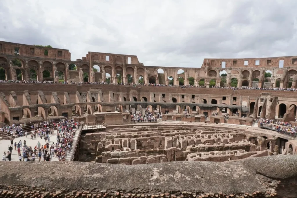 Vista interna e aérea do Coliseu de Roma, mostrando a arena em ruínas (hipogeu) e a multidão de turistas nas áreas de visitação dos andares superiores e inferiores em um dia parcialmente nublado.